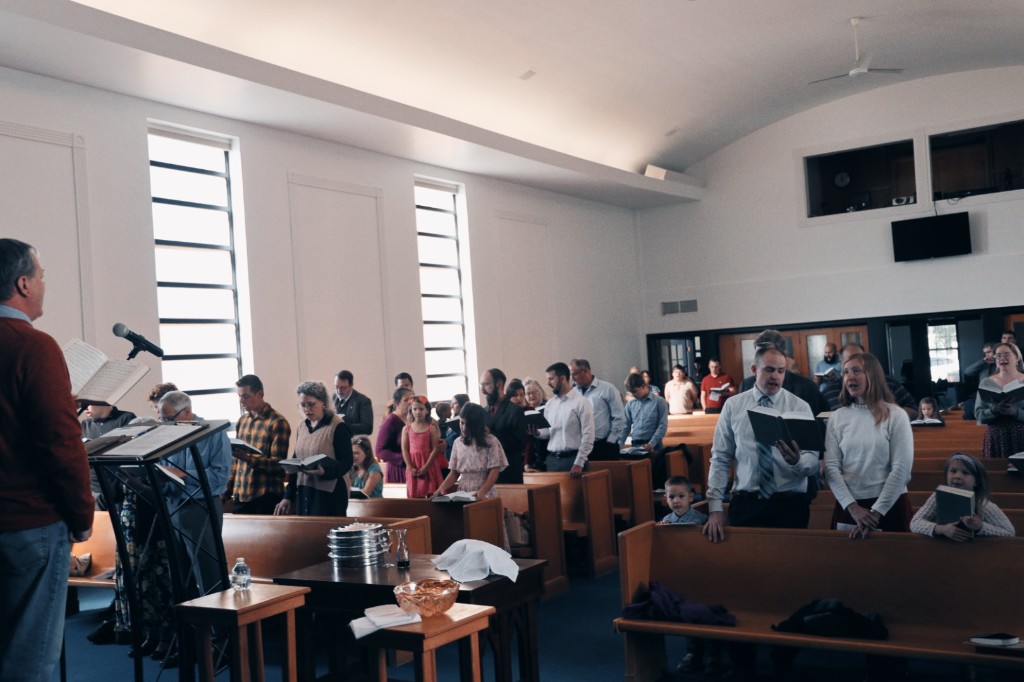 Sunday worship at Christ Church of Livingston County: a leader reads from the lectern with the Lord's Supper table set in the foreground while the congregation stands and sings